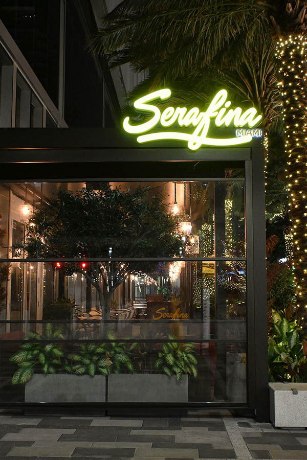 bright-yellow-neon-sign-for-Serafina-Miami-glows-above-a-glass-enclosed-patio-filled-with-lush-green-plants
