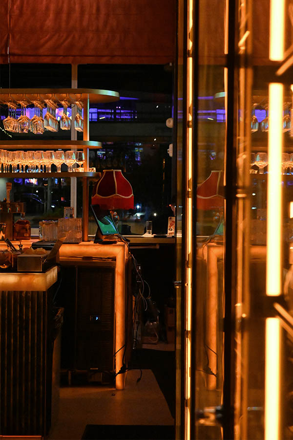 dimly-lit-bar-area-features-golden-shelves-stacked-with-gleaming-glassware-next-to-a-glowing-vertical-light-strip-and-a-small-red-lamp-with-fringe-shading-interior-design
