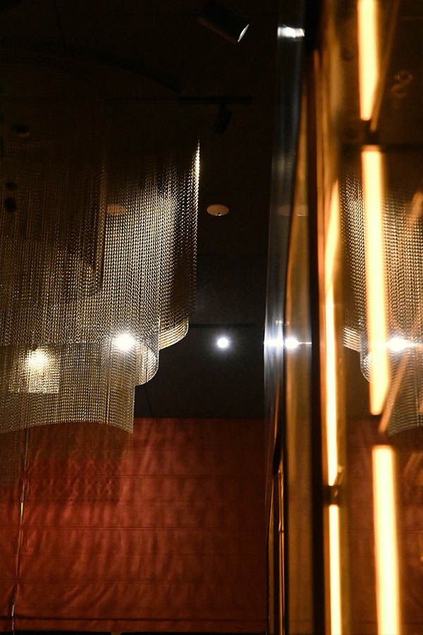 elegant-gold-chain-chandelier-cascades-downward-next-to-a-reflective-glass-surface-illuminated-by-glowing-vertical-strips-of-warm-light-serafine-miami