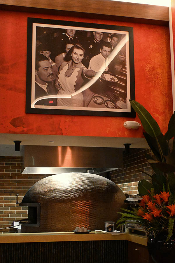 framed-vintage-photo-hangs-on-an-orange-wall-above-a-metallic-domed-pizza-oven-and-orange-flowers