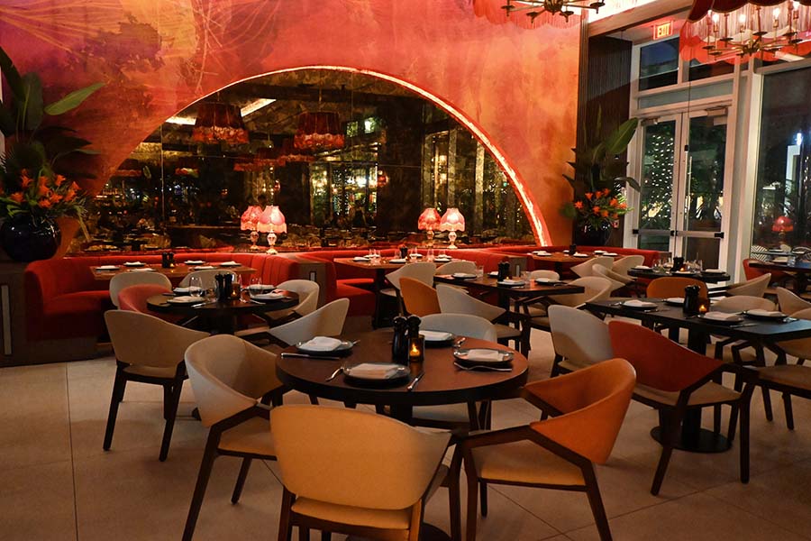 indoor-dining-room-features-numerous-round-wooden-tables-and-upholstered-chairs-arranged-before-a-large-arched-wall-decorated-with-vibrant-pink-and-orange-abstract-art